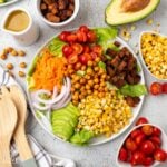 Vegan cobb salad on a white plate wiht an avocado and bowls of ingredients around it.