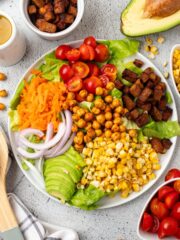 Vegan cobb salad on a white plate wiht an avocado and bowls of ingredients around it.