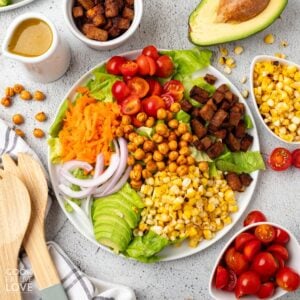 Vegan cobb salad on a white plate wiht an avocado and bowls of ingredients around it.
