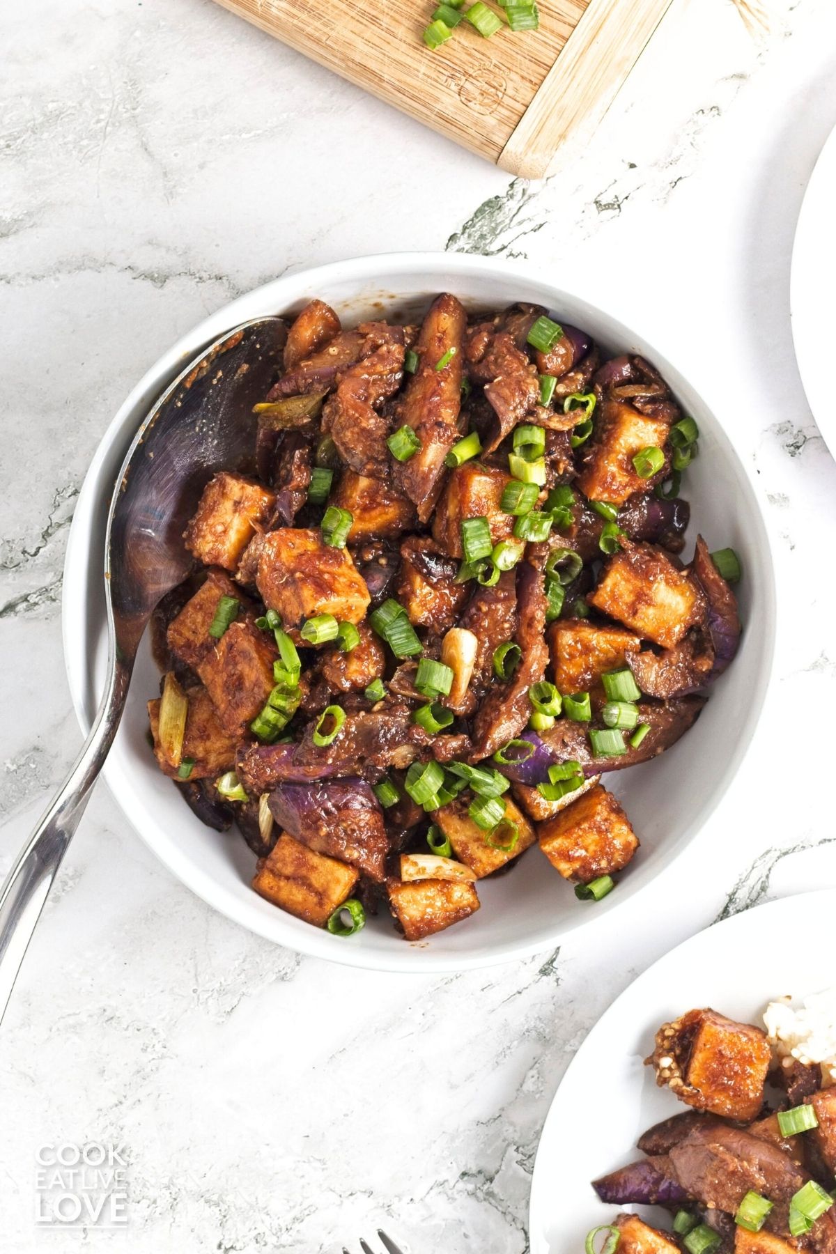 A bowl of tofu eggplant stir fry garnished with green onions and a spoon resting in a bowl.