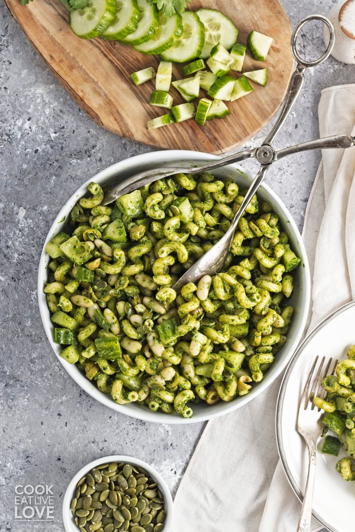 A bowl of green goddess pasta salad with diced cucumber on a board to the side.