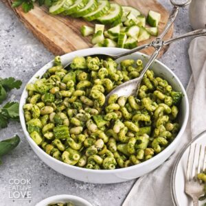 A bowl of green goddess pasta salad with sliced cucumbers in the background.