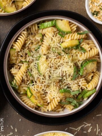 Lentil zucchini pasta in a bowl with bowls of garnishes around the edges of the bowl.