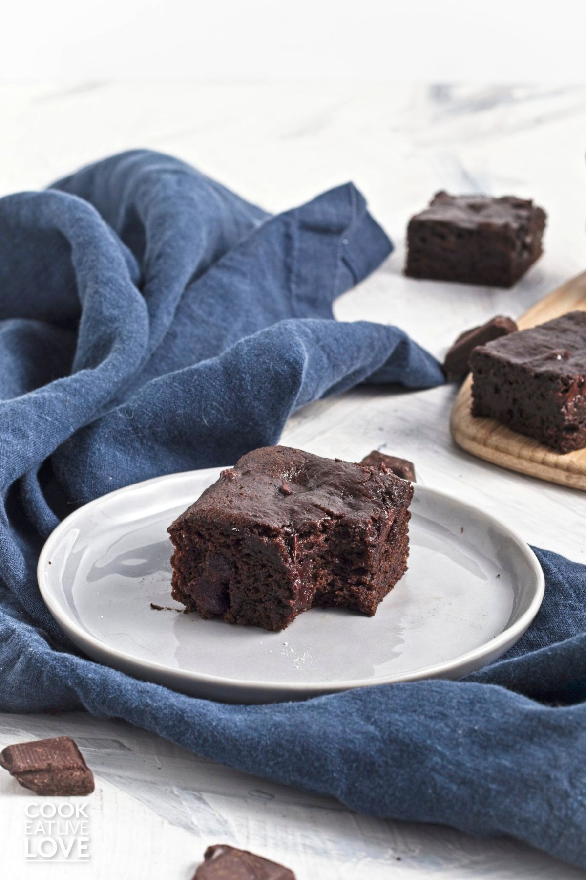 A zucchini brownie with a bite missing on a white plate set on top of a blue tea towel.