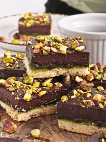 A stack of pistachio bars with chocolate ganache and chopped pistachios on a wooden board.