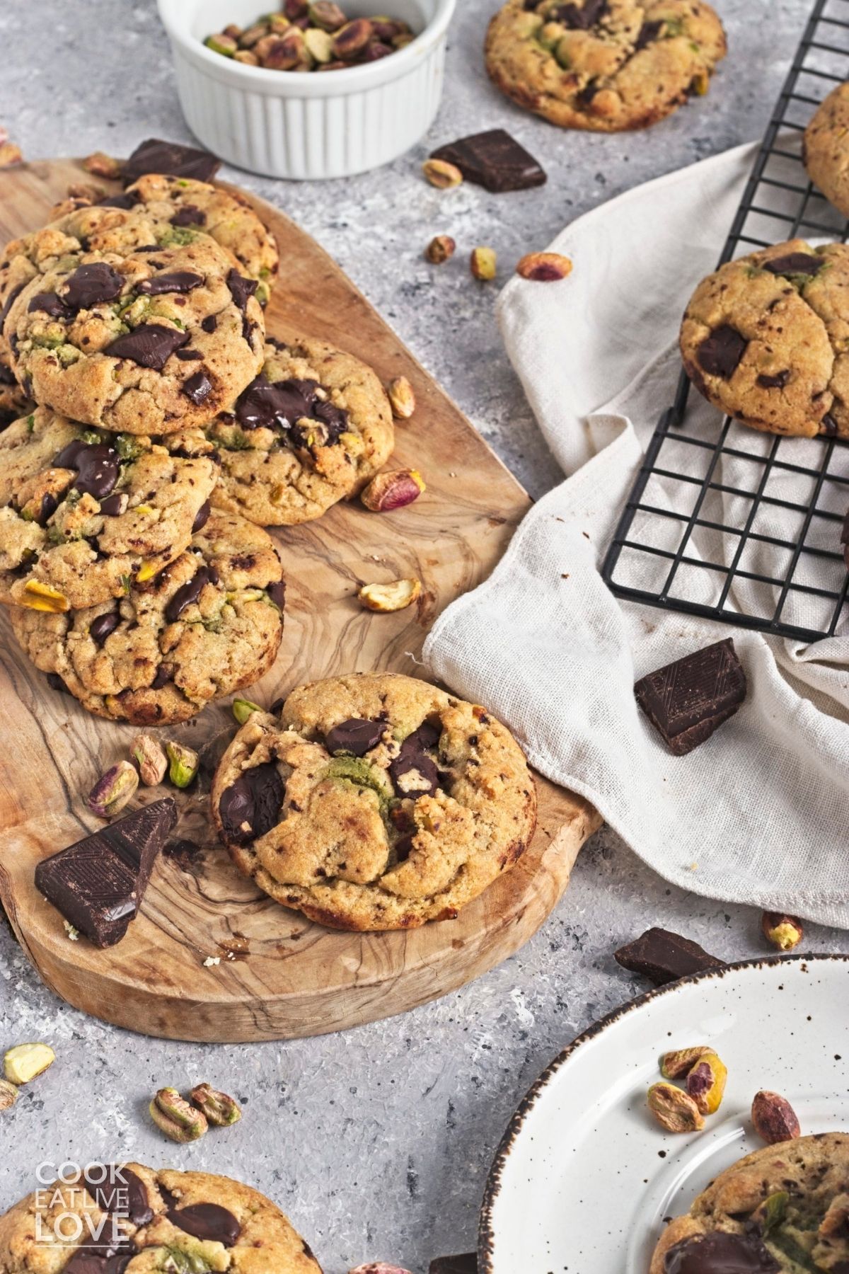 Pistachio chocolate chunk cookies on a wooden platter with more on a rack and a plate.