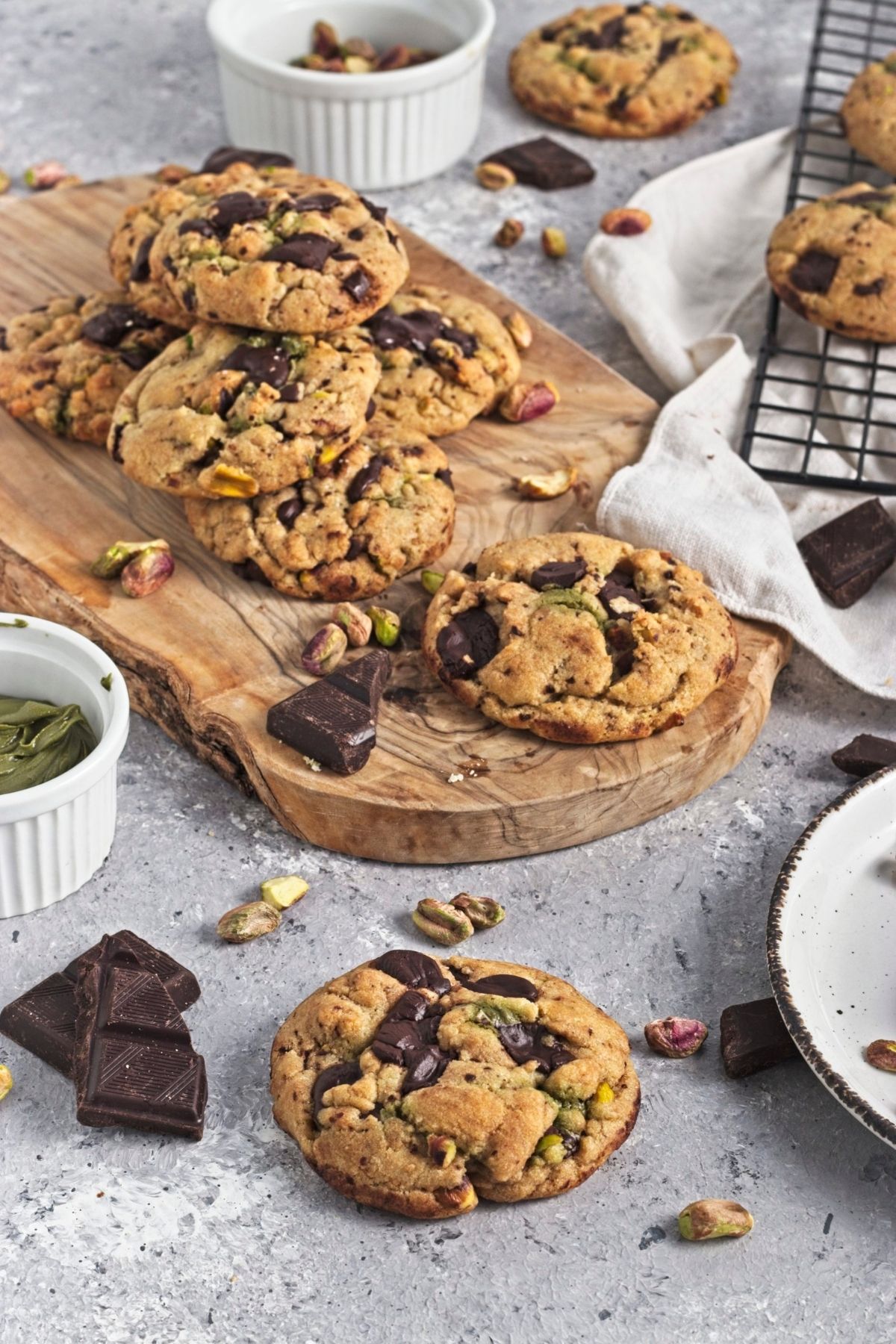 Pistachio chocolate chip cookies on a wooden board with nuts and chocolate chunks around.