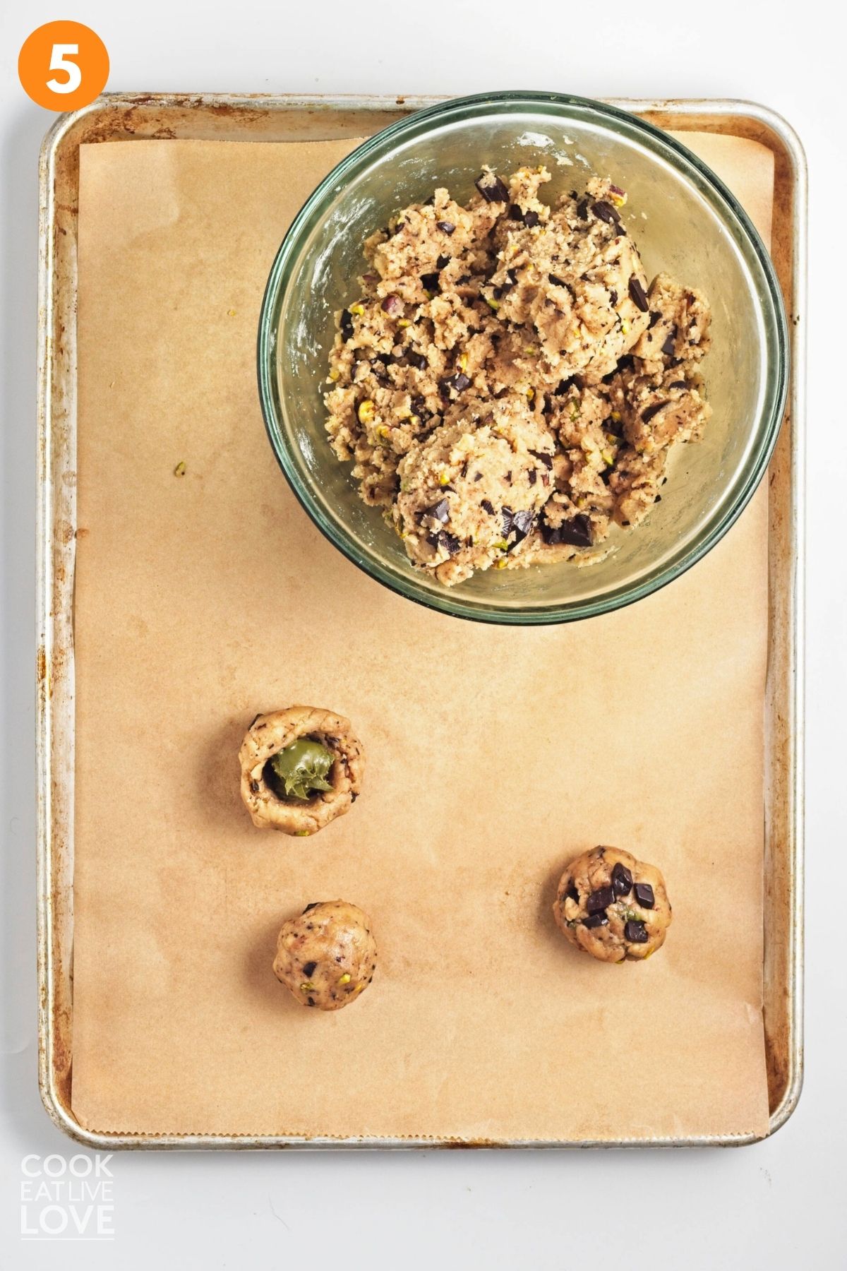 A bowl of cookie dough with some cookies rolled into a ball and one adding the pistachio cream to the inside.