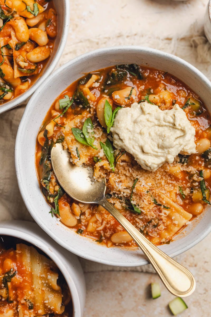 A bowl of vegan lasagna soup with a spoon resting in it and vegan ricotta on top.