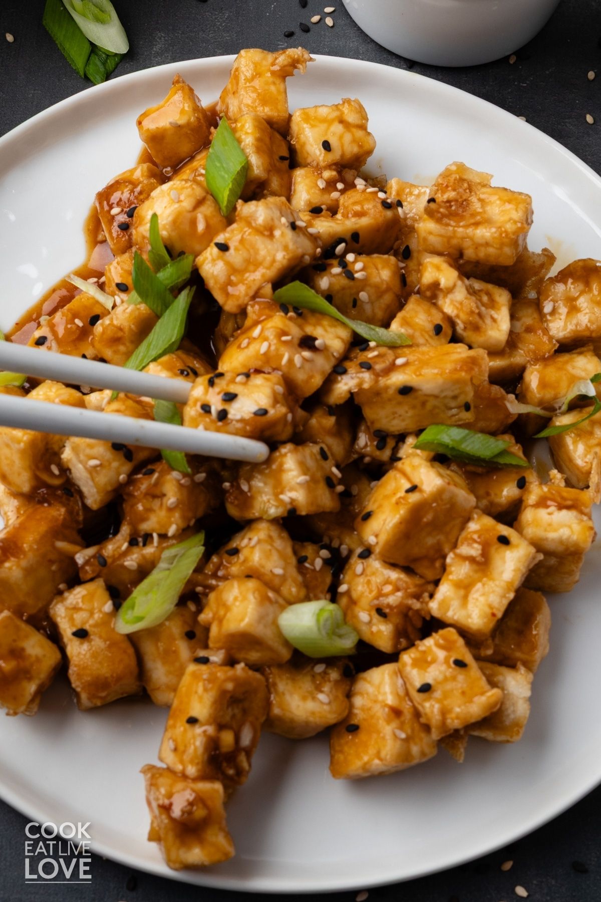 Chopsticks grabbing a piece of honey garlic tofu from a plate.