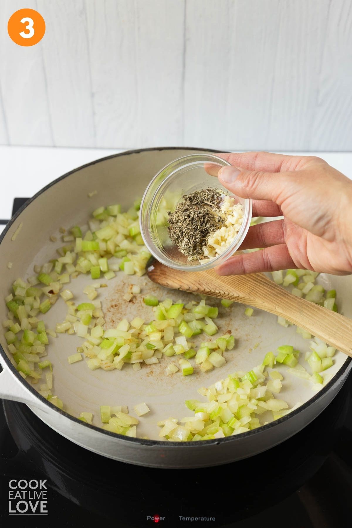 A hand holding seasoning over a skillet of cooked celery and onion. 