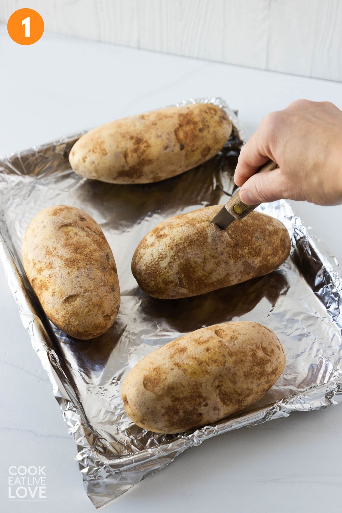 Potatoes on a baking tray with a hand holding  a fork to poke it with holes. 